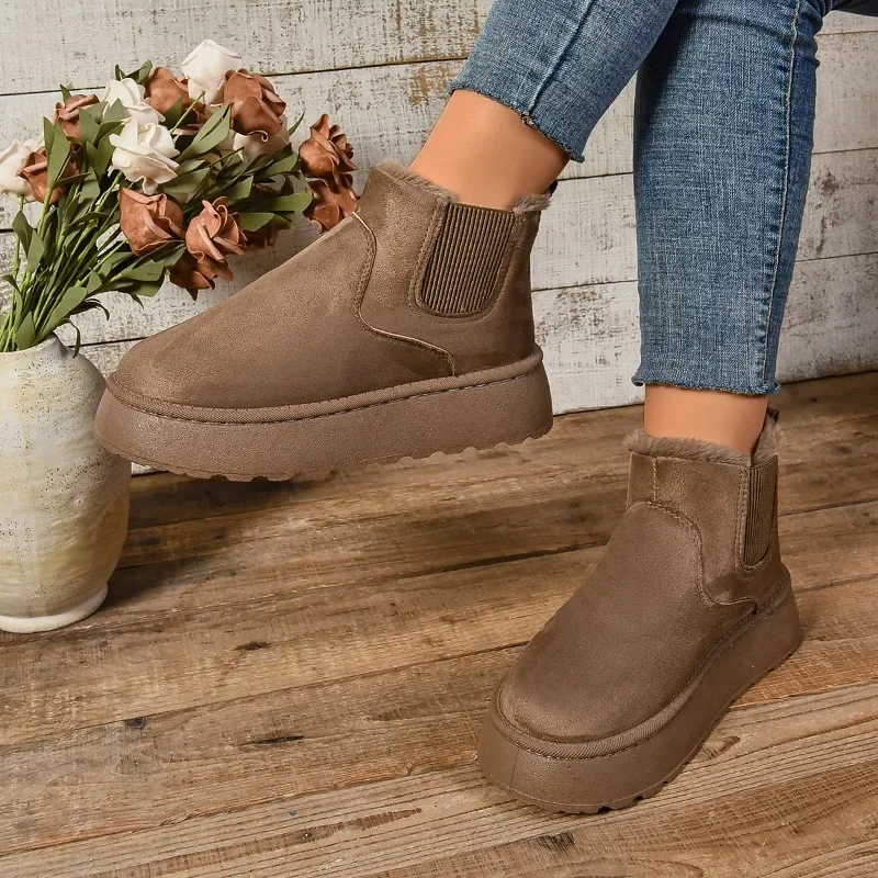 A pair of brown suede ankle boots with a platform sole. The boots are shown on a woman's feet, and they are sitting on a wooden floor.