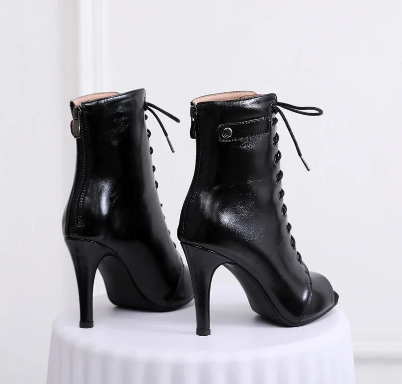 A pair of black peep-toe lace-up stiletto boots sits on a white pedestal.