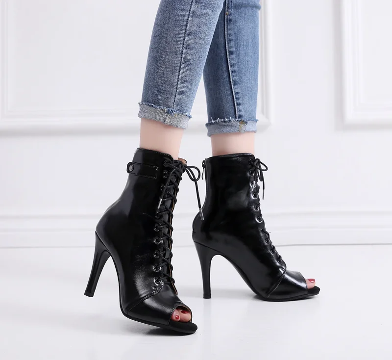 A pair of black peep-toe lace-up stiletto boots are shown on a model's feet. The model is wearing blue jeans.