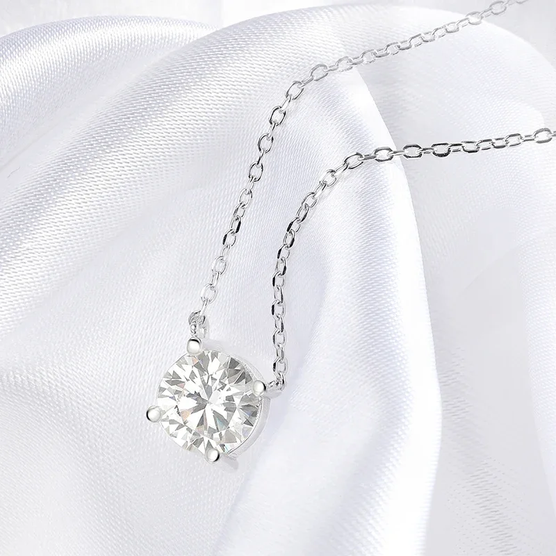 Close-up view of a solitaire diamond pendant necklace resting on white silk fabric. The necklace features a delicate chain and a single, brilliant-cut diamond.