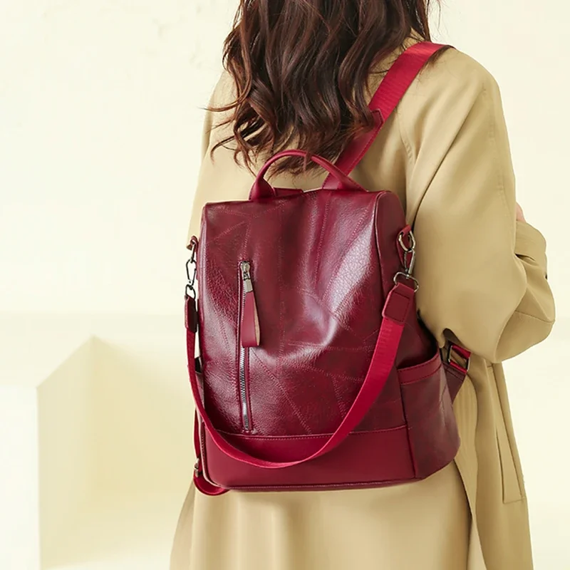 A maroon leather backpack with a red strap is worn by a model. The model is wearing a beige coat.