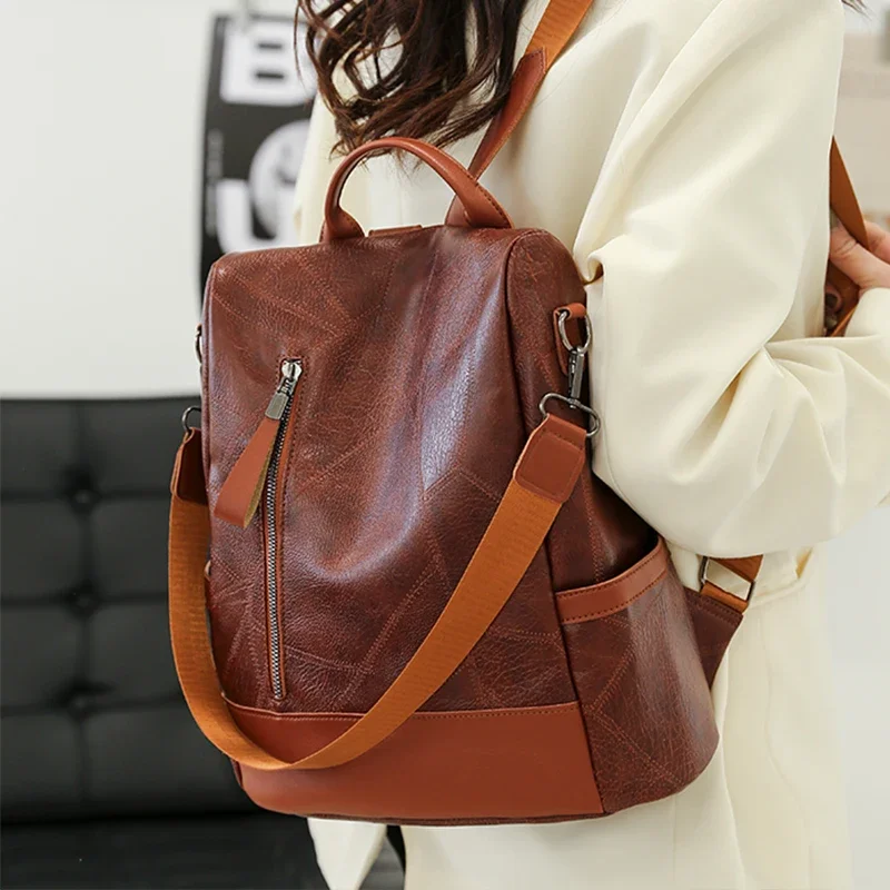 A brown leather backpack with orange straps is shown being worn by a person wearing a cream-colored sweater. The backpack is stylish and has a lot of pockets.